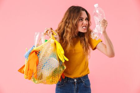 Photo of young furious woman holding bag with plastic waste and bottle camera isolated over pink...