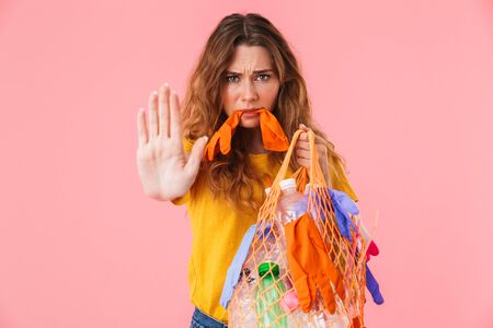 Photo of confused annoyed woman holding bag with plastic waste and showing stop gesture isolated...