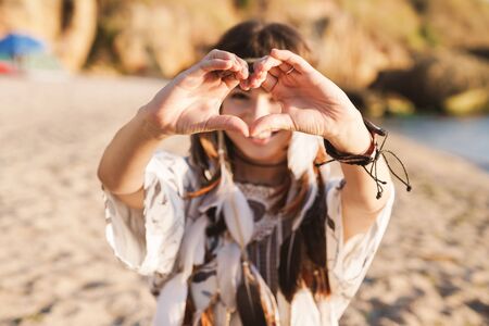Image of smiling hippy girl in feather headband showing heart shape while walking by seaside in morningの写真素材