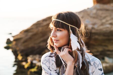 Image of young hippy girl wearing feather headband walking by seaside in morning