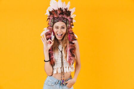 Portrait of delighted nice woman in headdress of feathers winking and gesturing ok sign isolated...