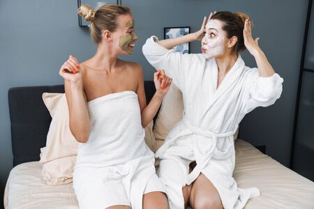 Image of a happy cheerful young women friends sisters at home in towel and bathrobe after spa procedure taking care of their skin with face mask sit on bed.の写真素材