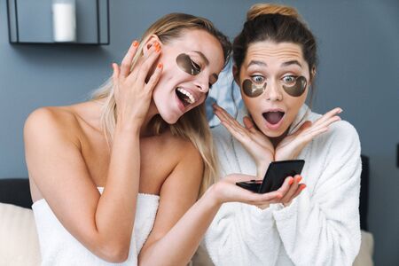 Photo of a positive shocked young women friends sisters indoors at home in towel and bathrobe after spa procedure holding mirror taking care of their skin with under eye patches.の写真素材