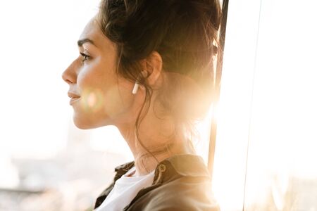 Image of lovely brunette woman listening to music with earpods while standing at terrace outdoors in morningの写真素材