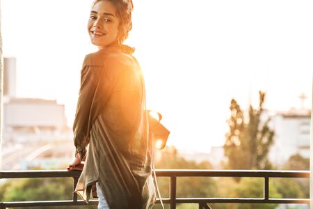 Image of happy brunette smiling woman carrying backpack standing in front of amazing sunrise over city while walking outdoorsの写真素材