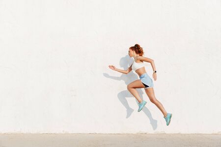 Image of redhead young woman in sportswear running along white wall while doing workout in morningの写真素材