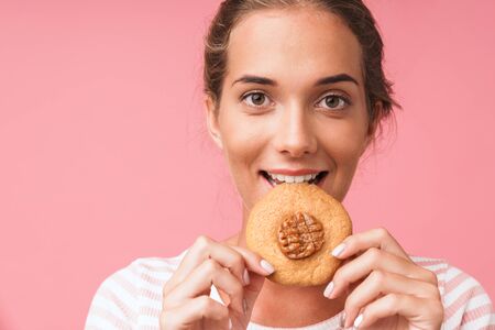 Image closeup of caucasian gorgeous woman smiling and eating tasty cookie isolated over pink...