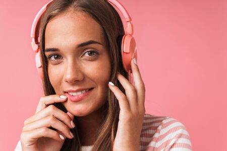 Image closeup of charming beautiful girl wearing striped clothes smiling and listening to music with headphones isolated over pink backgroundの写真素材
