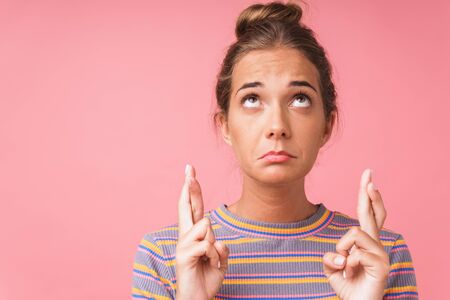 Image closeup of sad caucasian woman wearing striped t-shirt looking upward with fingers crossed isolated over pink wallの写真素材