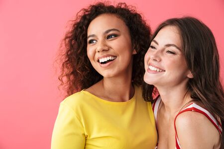 Image of cheerful multinational girls smiling and looking aside at copyspace isolated over pink...