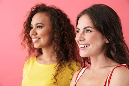 Image of satisfied multinational girls smiling and looking aside at copyspace isolated over pink backgroundの写真素材
