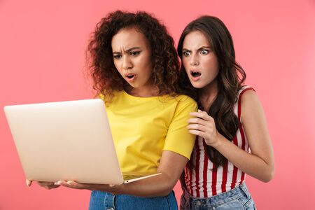 Image of shocked multinational girls wondering and holding laptop computer isolated over pink backgroundの写真素材
