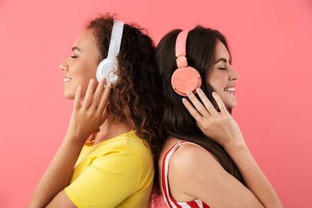 Image of charming multinational girls standing back to back and listening to music with headphones isolated over pink backgroundの写真素材
