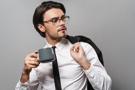 Portrait of a handsome serious young businessman wearing suit standing isolated over gray background, drinking coffee, looking awayの写真素材