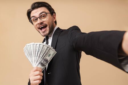 Portrait of a handsome young businessman wearing suit standing isolated over beige background, showing money banknotes while taking a selfieの写真素材