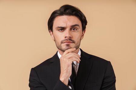 Portrait of a handsome pensive young businessman wearing suit standing isolated over beige background, thinkingの写真素材