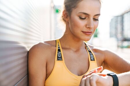 Image of caucasian woman in sportswear looking at smartwatch while walking outdoors in sunny morning