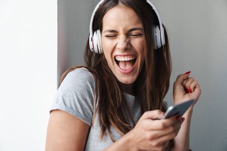 Close up of a young smiling beautiful girl standing leaning on a gray wall over gray background, listening to music with wireless headphones while holding mobile phone, singingの写真素材