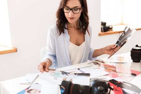 Image of a concentrated young photographer woman in office sit at the table holding photos.の写真素材