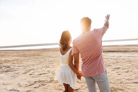 Back view of a beautiful young couple wearing summer clothing standing at the beach, pointing finger awayの写真素材