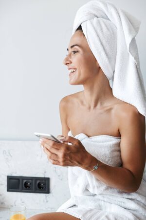 Photo of a young positive woman in towel after spa procedure shower indoors at home at the kitchen using mobile phone.の写真素材