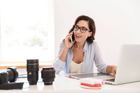 Image of a smiling optimistic young photographer woman in office sit at the table with camera lenses...