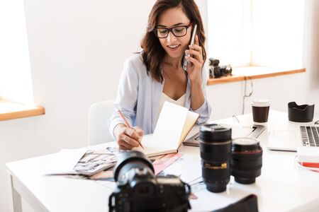 Photo of a smiling young woman photographer talking by mobile phone using laptop computer writing notes.の写真素材
