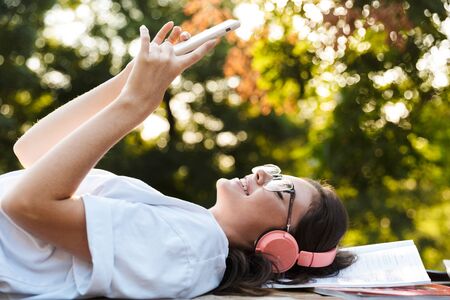 Image of a smiling happy student lady outdoors in nature green park lies on grass listening music with headphones on copybooks using mobile phone.の写真素材