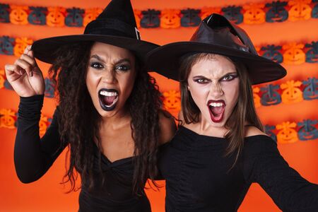 Image of frightening witch women in black halloween costume and makeup screaming isolated over orange pumpkin wallの写真素材