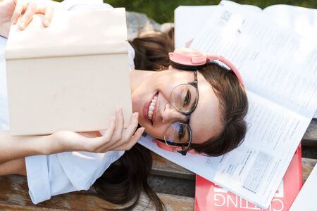 Photo of a cheery happy smiling student lady outdoors in nature green park lies on grass listening...