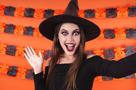Image of amazed witch girl in black halloween costume smiling and waving hand at camera isolated...