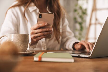 Cropped photo of young business woman sit indoors in office using laptop computer and mobile phone.の写真素材