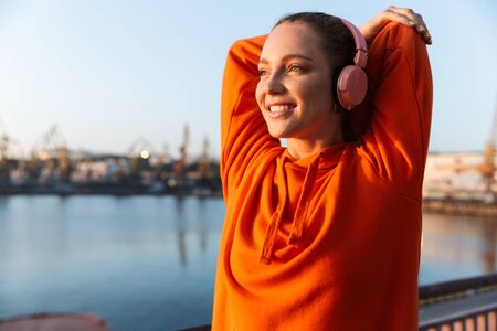 Attractive young fitness woman wearing hoodie standing at the beach, listening to music with headphones, doing stretching exercisesの写真素材