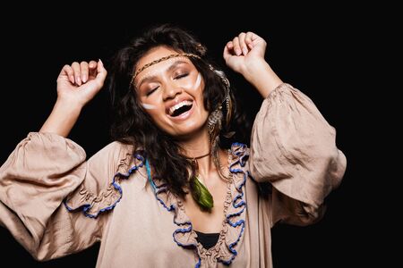 Portrait of a beautiful cheerful young brunette woman wearing tribal clothing and accessories standing isolated over black background, dancingの写真素材