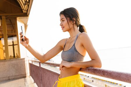 Image of beautiful asian woman in sportive clothes taking selfie photo on smartphone while standing over railing at sea portの写真素材