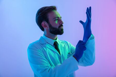 Portrait of a confident young man doctor wearing uniform standing isolated over pink-blue haze background, putting gloves onの写真素材