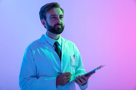 Portrait of a confident young man doctor wearing uniform standing isolated over pink-blue haze background, taking notes in a notepadの写真素材