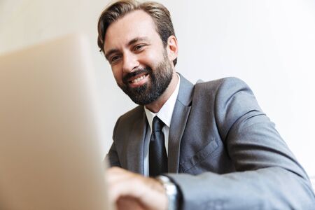 Image of a handsome cheerful happy young positive business man sitting in office using laptop computer working indoors.の写真素材