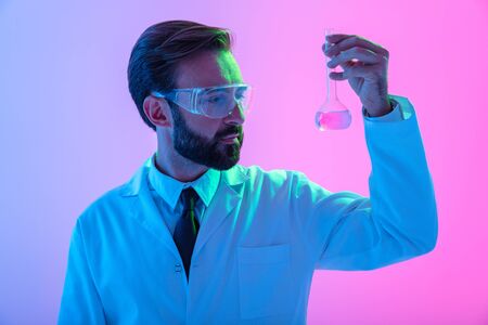 Portrait of a confident man scientist wearing unifrom and glasses standing isolated over blue-pink haze background? looking at a flask with liquidの写真素材