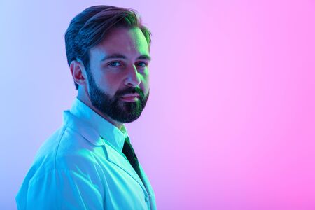 Portrait of a confident young man doctor wearing uniform standing isolated over pink-blue haze background, looking at cameraの写真素材