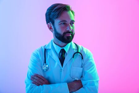 Portrait of a confident young man doctor wearing uniform standing isolated over pink-blue haze background, arms folded, looking awayの写真素材