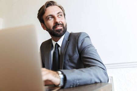 Portrait of a handsome positive bearded business man sitting in office using laptop computer working indoors looking aside.の写真素材