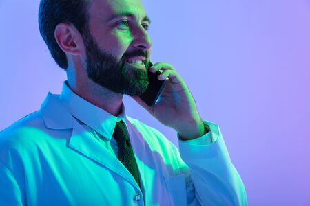 Portrait of a smiling young man doctor wearing uniform standing isolated over pink-blue haze background, talking on mobile phoneの写真素材