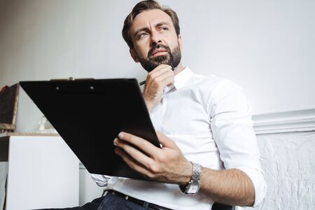 Image of puzzled concentrated businessman wearing wristwatch holding clipboard and looking aside...