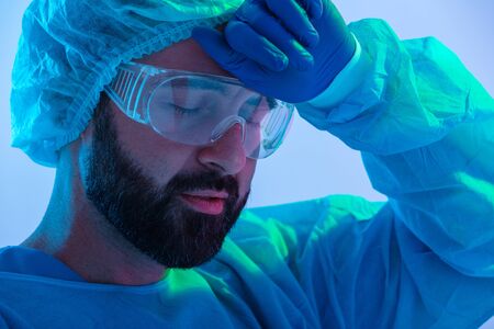 Portrait of a tired man scientist wearing unifrom and glasses standing isolated over blue-pink haze background, wiping foreheadの写真素材