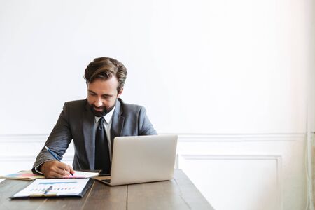 Image of unshaven masculine businessman wearing formal suit working at laptop in office while writing down notesの写真素材