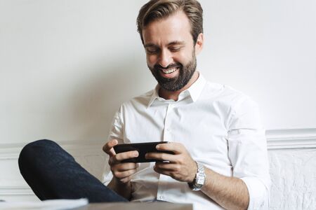 Image of successful laughing businessman wearing white shirt playing video game on cellphone while sitting at table in officeの写真素材