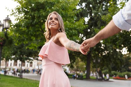Image of a pleased optimistic cute young loving couple walking by street outdoors holding hands of each other.の写真素材