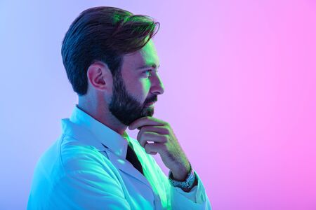 Portrait of a confident young man doctor wearing uniform standing isolated over pink-blue haze background, looking awayの写真素材