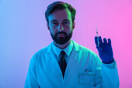 Portrait of a confident young man doctor wearing uniform standing isolated over pink-blue haze...